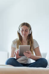 Frau mit Tablet-PC, die über Kopfhörer Musik hört, auf dem Bett sitzend - PGF01074