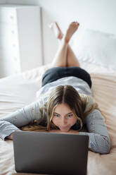 Lächelnde Frau mit Laptop auf dem Bett liegend zu Hause - PGF01076