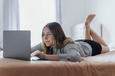 Lächelnde Frau mit Laptop auf dem Bett liegend zu Hause - PGF01079
