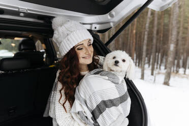 Lächelnde Frau mit niedlichem Hund, eingewickelt in eine Decke, sitzt im Kofferraum eines Autos - SIF00071