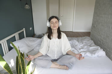 Frau mit drahtlosen Kopfhörern beim Meditieren auf dem Bett zu Hause - VBUF00087