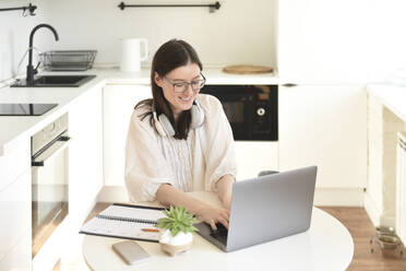 Happy freelancer using laptop sitting at table in kitchen - VBUF00111
