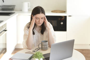 Stressed freelancer with laptop sitting at table in kitchen - VBUF00112