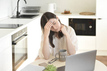 Stressed freelancer with laptop sitting at table in kitchen - VBUF00113