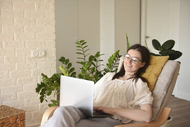 Glückliche Frau mit Laptop auf einem Stuhl zu Hause sitzend - VBUF00118