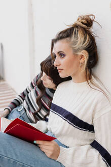 Junge Frau mit Buch, die neben einem Freund sitzt, der sich mit dem Kopf auf die Schulter vor einer Wand stützt - MRRF01969