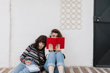Woman peeking from behind book by friend leaning head on shoulder in front of white wall - MRRF01971