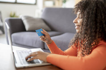 Smiling woman holding credit card sitting with laptop at home - JCZF01006