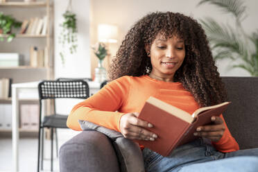 Young woman reading book sitting on sofa in living room - JCZF01008