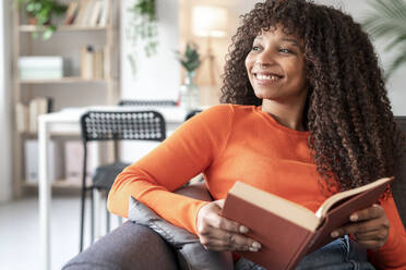 Glückliche Frau mit lockigem Haar, die ein Buch hält und zu Hause auf dem Sofa sitzt - JCZF01009