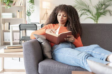 Young woman with curly hair reading book sitting on sofa at home - JCZF01011