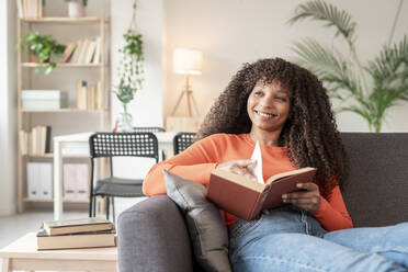 Smiling woman with book sitting on sofa in living room - JCZF01013