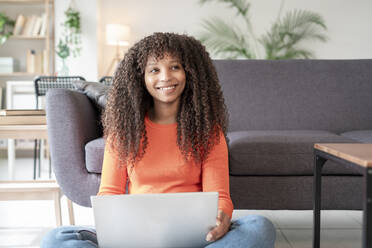 Glückliche Frau mit Laptop, die zu Hause vor dem Sofa sitzt - JCZF01014