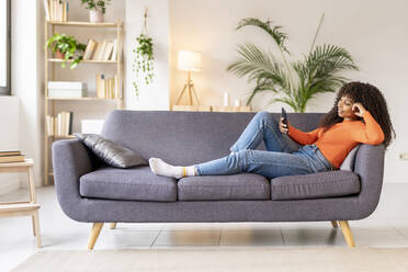 Young woman using smart phone sitting on sofa in living room - JCZF01015