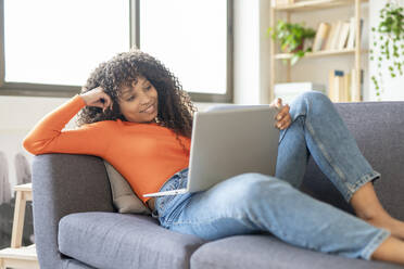 Smiling young woman using laptop sitting on sofa at home - JCZF01017