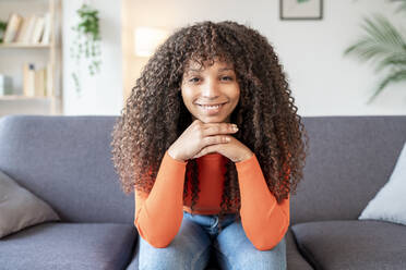 Smiling young woman with curly hair sitting on sofa in living room - JCZF01021