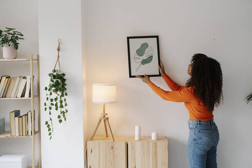 Junge Frau mit lockigem Haar hängt Bilderrahmen an der Wand zu Hause - JCZF01026