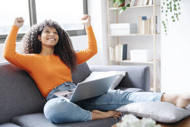 Happy woman gesturing fist sitting with laptop on sofa at home - JCZF01029