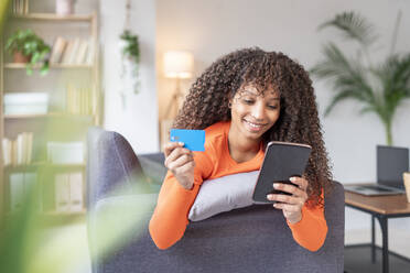 Smiling woman holding credit card using mobile phone at home - JCZF01033