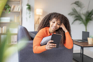Lächelnde Frau mit lockigem Haar, die auf dem Sofa zu Hause ein Mobiltelefon benutzt - JCZF01034