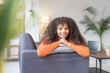 Lächelnde Frau mit lockigem Haar, die sich auf dem Sofa im Wohnzimmer anlehnt - JCZF01036