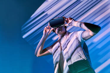 Mann in modischem Hemd und moderner VR-Brille erlebt virtuelle Realität, während er in der Nähe einer Wand mit leuchtenden Linien im Studio steht - ADSF34528