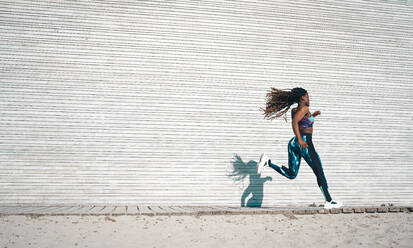 Full body side view of slim African American female with flying long dreadlocks wearing tight sportswear jogging on walkway near metal wall during cardio workout - ADSF34547
