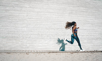 Ganzkörper-Seitenansicht einer schlanken afroamerikanischen Frau mit fliegenden langen Dreadlocks in enger Sportkleidung, die auf einem Gehweg in der Nähe einer Metallwand während eines Cardio-Trainings joggt - ADSF34547