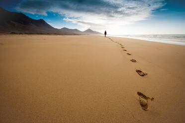 Ein unerkannter Reisender hinterlässt Fußabdrücke im Sand, während er am Strand spazieren geht und die Freiheit im Urlaub auf der hügeligen Insel Fuerteventura genießt - ADSF34548