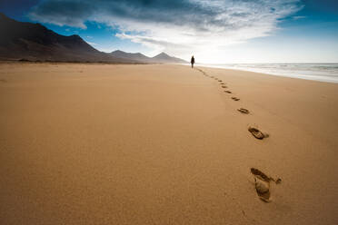Unrecognizable traveler leaving footprints on sand while strolling on shore and enjoying freedom during vacation on hilly Fuerteventura island - ADSF34548