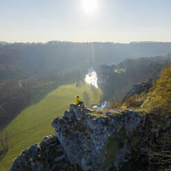 Aerial view of a woman sitting on the rocks with Walzin castle in background at sunset, Dinant, Namur, Belgium. - AAEF14439