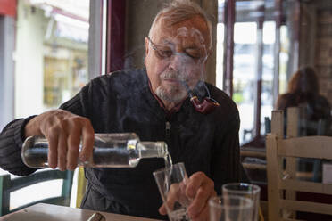 Älterer Mann, der Zigarre raucht und ein Getränk in ein Glas gießt, sitzt am Tisch - MAMF02193