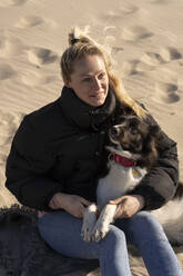 Lächelnde junge blonde Frau sitzt mit Hund im Sand - SSGF00725