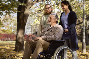 Smiling senior woman talking with man sitting in wheelchair pushed by nurse at park - ABIF01650