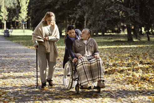 Ein behinderter älterer Mann unterhält sich mit einer Frau, die mit einem Stock geht, der von einem Pfleger geschoben wird, der einen Rollstuhl im Park schiebt - ABIF01653