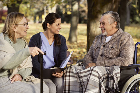 Lächelnde Krankenschwester bespricht Dokumente mit einem älteren Ehepaar im Park - ABIF01659
