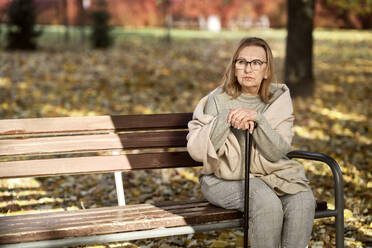 Ältere Frau mit Brille sitzt auf einer Bank mit Spazierstock im Park - ABIF01664