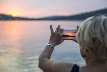 Ältere Frau fotografiert Sonnenuntergang mit dem Smartphone im Urlaub - MAMF02201