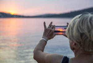 Senior woman photographing sunset through smart phone on vacations - MAMF02201