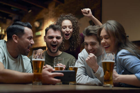 Group of cheerful soccer fans having beer and watching a match on smartphone in a pub - ABIF01684