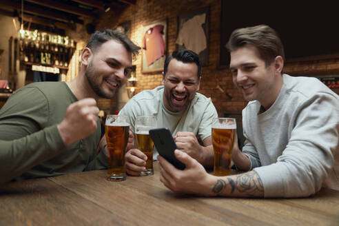 Drei fröhliche männliche Fußballfans, die in einer Kneipe Bier trinken und ein Spiel auf dem Smartphone verfolgen - ABIF01686