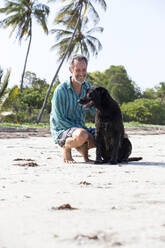 Lächelnder Mann hockt mit Hund am Strand an einem sonnigen Tag - NDF01416