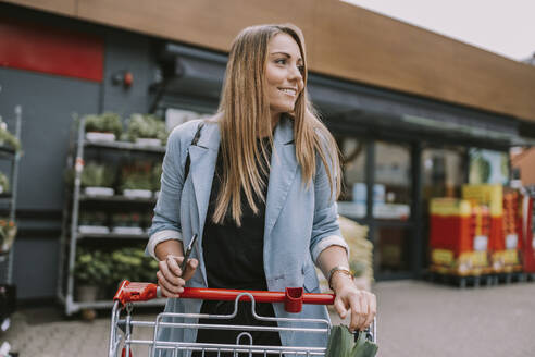 Lächelnde Frau mit Smartphone und Einkaufswagen vor einem Geschäft stehend - MFF08991