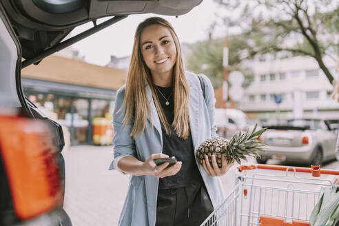 Lächelnde Frau mit Smartphone und Ananasfrucht am Kofferraum eines Autos - MFF08995