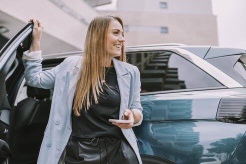 Glückliche Frau mit Mobiltelefon an der Autotür stehend - MFF08999