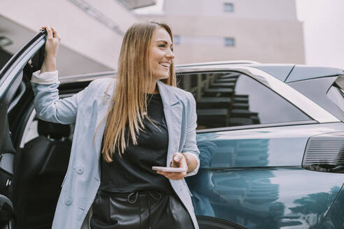 Happy woman with mobile phone standing by car door - MFF08999