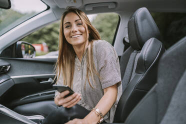 Glückliche blonde Frau mit Smartphone sitzt auf dem Beifahrersitz im Auto - MFF09002
