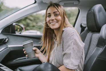 Smiling woman with disposable coffee cup sitting in car - MFF09003