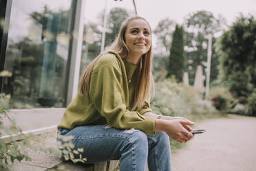 Thoughtful young woman with smart phone sitting on wall - MFF09020