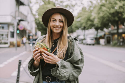 Happy young woman with sandwich standing on street - MFF09027